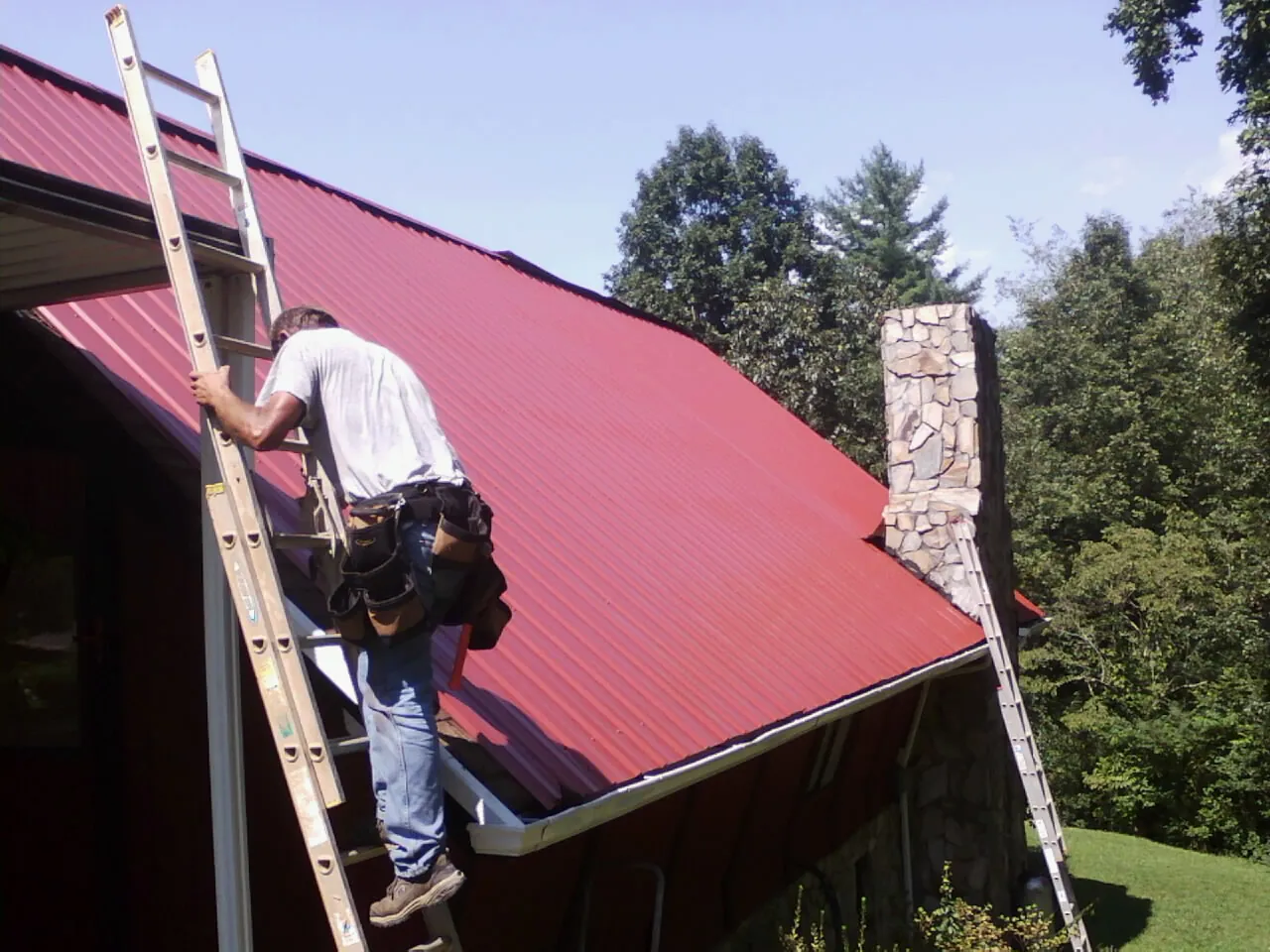 Roofer on a ladder beside a red metal roof and stone chimney — Soffit & Fascia Repair by Roofing Brothers in Blountville